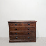 A rustic dark brown wooden chest of drawers with five drawers and round black knobs, set against a plain off-white wall and concrete floor.