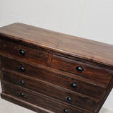 A dark brown wooden dresser with five drawers and round black knobs, photographed against a plain off-white wall.