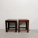 A pair of dark brown wooden stools with woven straw seats, positioned side-by-side against a plain white wall and concrete floor.