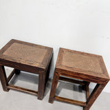 A pair of rustic dark brown wooden stools with woven cane seats, photographed against a plain white wall and light gray floor.