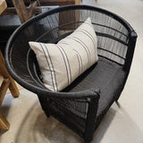 A black woven outdoor armchair with a cream throw pillow featuring black stripes, set in a rustic furniture showroom.