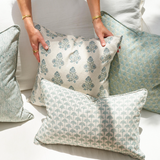 Three decorative throw pillows with green and beige botanical block-print patterns are arranged on a white sofa, with a person's hands holding one pillow.