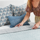 A woman is shown from the side as she styles a bed with patterned pillows and a textured light blue and white duvet cover. The bedding features a blue and white floral block print on the pillows and a matching abstract pattern on the duvet, with a textured gray throw blanket partially folded.