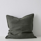 A square throw pillow in a textured olive green fabric, leaning against a white wall on a white surface.