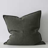 A square throw pillow in a dark olive green fabric with contrasting dark green piped edges, resting against a light gray textured wall.