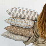 A person holding a stack of four decorative throw pillows with various geometric and bohemian patterns in brown, beige, and gray tones, set against a white paneled wall.