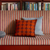 A cozy indoor seating area with a built-in bench upholstered in red, orange, and cream vertical stripes, set against a dark wood bookshelf. The bench is styled with three throw pillows: a gray round cushion, a bright orange patterned square cushion, and a light blue rectangular cushion.